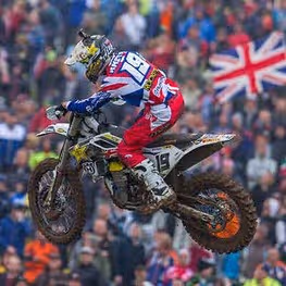 Motocross rider in red, white, and blue gear jumping on a dirt bike with the number 19, with a crowd and a British flag in the background.